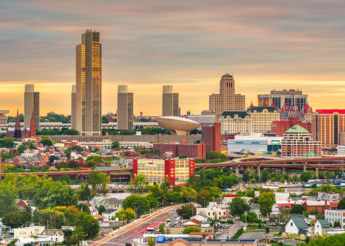 An aerial view of the Albany cityscape, with several skyscrapers, buildings, highways, and trees under an orange and yellow sunset.