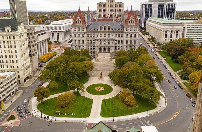 A birds-eye-view of the New York State Capitol, a large building with red spires on each corner and a bronze statue in front of a wide staircase along a circular pathway that surrounds a circle of grass. The building is in the center of a four-lane road, where there are dozens of cars parked along the sidewalk on the right side, and several buildings on each side.