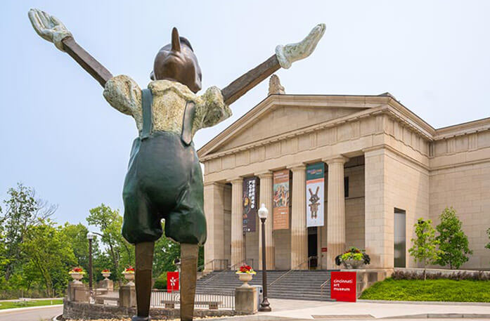 A large Pinocchio sculpture with his arms outstretched looking up to the sky stands in front of the Cincinnati Art Museum, a light brick building with four pillars and several stairs leading up to the door.