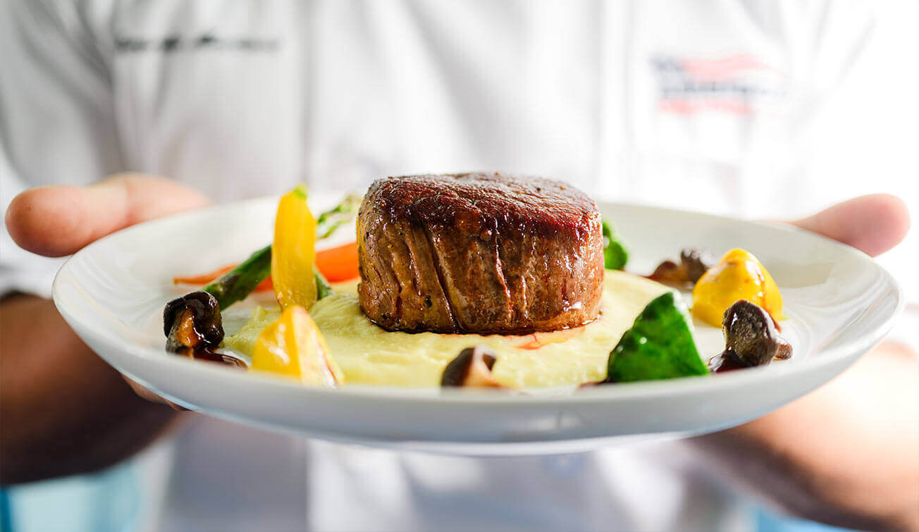 A chef presenting a plate with a filet mignon on mashed potatoes and other vegetables