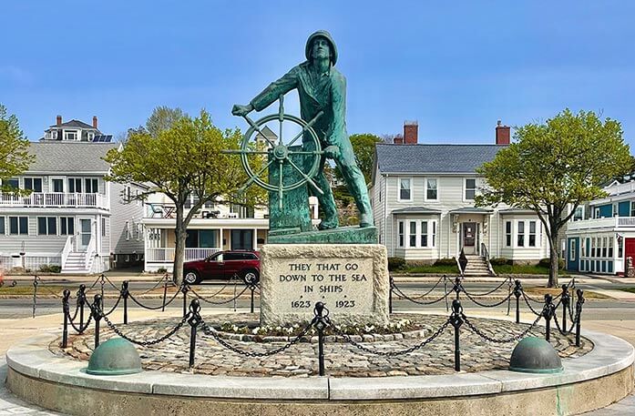 Gloucester Fisherman's Memorial, a large, green statue of a fisherman standing and holding onto the wheel of a ship in Gloucester, with a large stone stand below it with the phrase "They that go down to the sea in ships" and the years "1623" and "1923" on it. There is a circular platform made of stone with black railings surrounding the statue, and four houses behind it.