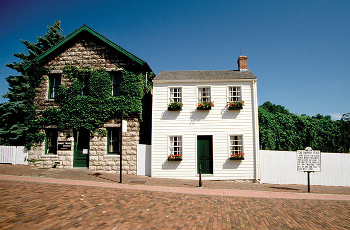 Mark Twain's light cream colored house with a dark green door and window planters to the right of a brown brick giftshop covered with green vines and topped with a green roof surrounded by the whitewashed fence of Tom Sawyer.