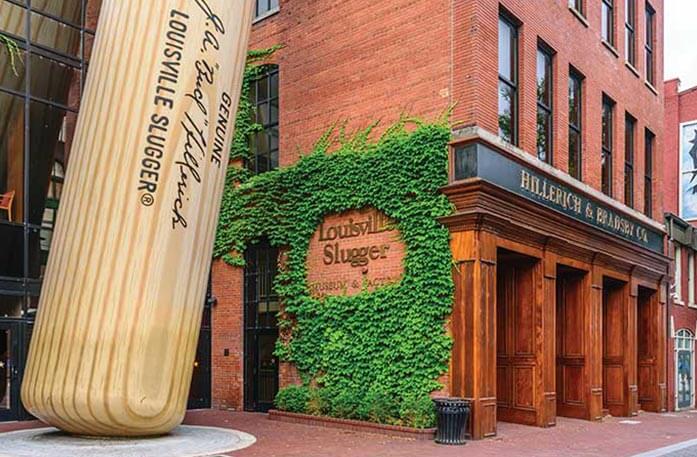 A giant wooden Louisville Slugger baseball bat leans against a red brick building, Hillerich & Bradsby Co., with "Louisville Slugger" in gold writing on the left side of the building surrounded by bright green vines in Louisville.