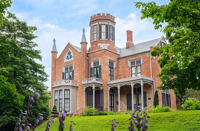 The Castle Historic House Museum in Marietta, a victorian-style brick mansion with small, ornate turrets and large windows surrounded by greenery and purple flowers.