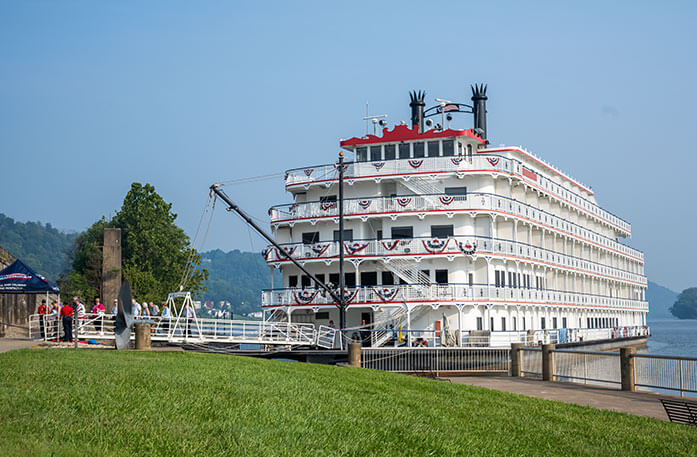 American Heritage paddlewheeler docked letting passengers off from the front of the white ship. It is decorated with patriotic red white and blue bunting flags on every railing.