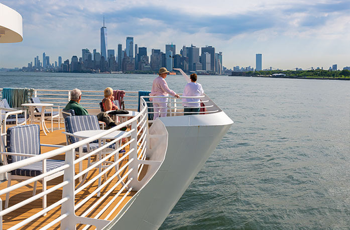 American Cruise Lines guests standing and sitting on the sun deck aboard a Coastal Cat with views of the New York City skyscrapers in the distance.