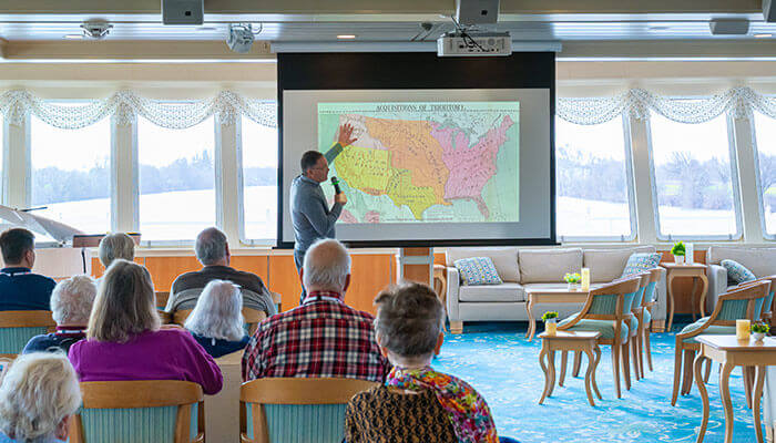 A presenter discussing a map image on the screen, in a lounge filled with cruise guests