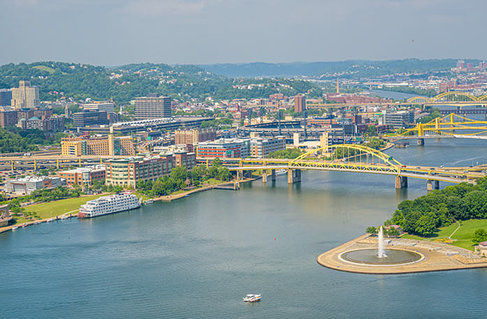 An aerial view of the Ohio River, containing a cityscape with mountains in the distance, several yellow bridges over the river, and American Heritage paddlewheeler docked along the river.