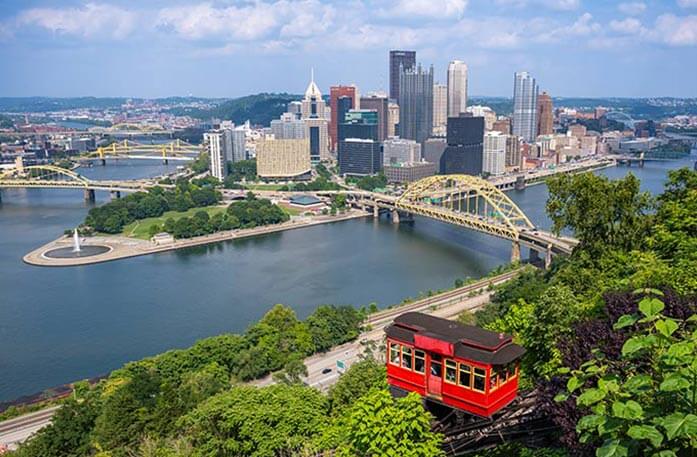 A red cable car descends a forest hill overlooking the Ohio River and a Pittsburgh cityscape featuring a brideg with a yellow truss in the middle and several skyscrapers under a partly cloudy sky.