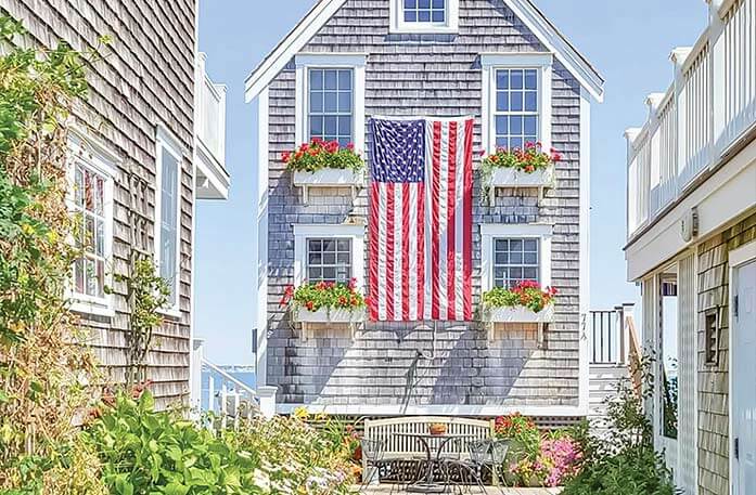 House in Provincetown, Massachusetts, with typical cape code shingles, four windows with windowboxes filled with red flowers, and in the center, the American Flag hanging down