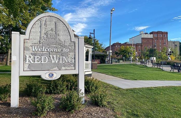 A grey welcome sign reads "Welcome to Red Wing" and is surrounded by brown mulch and small green plants with a red brick building and black benches in the background.