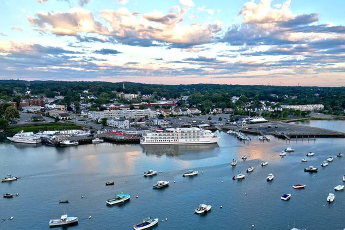 An aerial view of an American Cruise Lines Coastal Cat docked in Plymouth among several small boats on the water. On the shore, there are several buildings and thick trees under a cloudy sunrise.