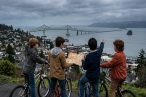 Four young boys on bicycles on a very gray day looking from a hilltop over the Columbia River, with two of them holding a map.