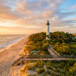 A white lighthouse rises above a palm-covered coastal island at sunset, with a wooden boardwalk crossing sandy dunes and a calm beach beside a marina.