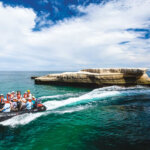 Cruise guests ride a rigid inflatable boat through turquoise sea beside dramatic limestone cliffs and a flat rock island under a bright blue sky.