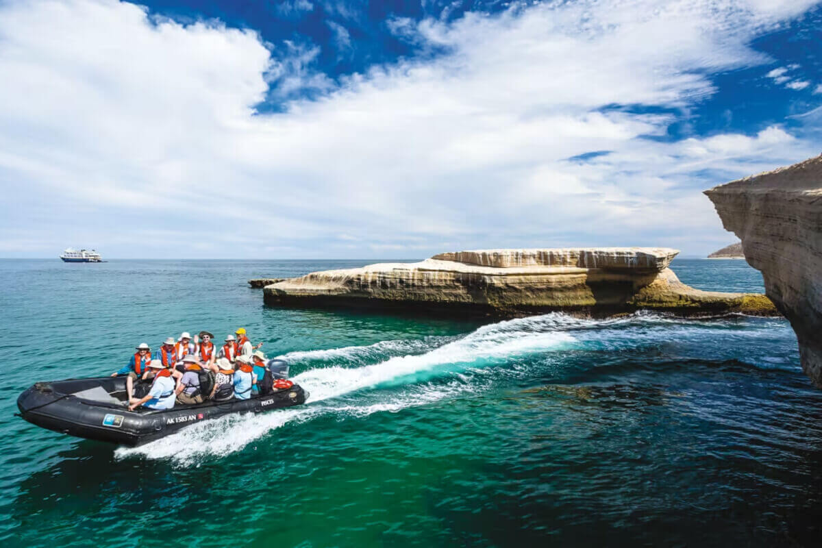 Cruise guests ride a rigid inflatable boat through turquoise sea beside dramatic limestone cliffs and a flat rock island under a bright blue sky.