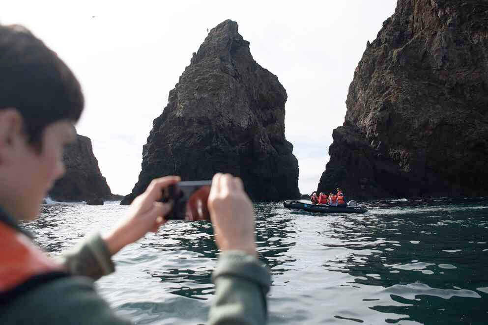 A cruise guest on a zodiac taking a photo of another zodiac filled with guests with a rocky Baja outcropping.
