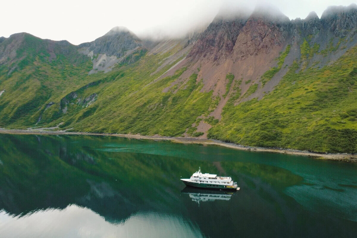An UnCruise ship glides across a calm emerald lake, reflecting steep green mountains veiled in mist beneath an overcast sky, creating a serene mood.