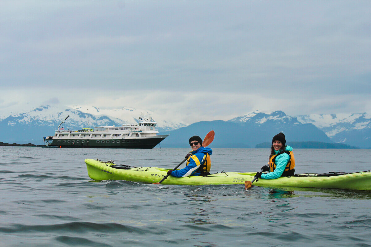 Two kayakers in bright gear paddle on calm water near a cruise ship, with snow-capped mountains under a cloudy sky; adventurous mood.