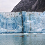 Towering blue glacier meets calm gray water, with tiny kayaks below and dark mountain cliffs behind, creating a dramatic scene.