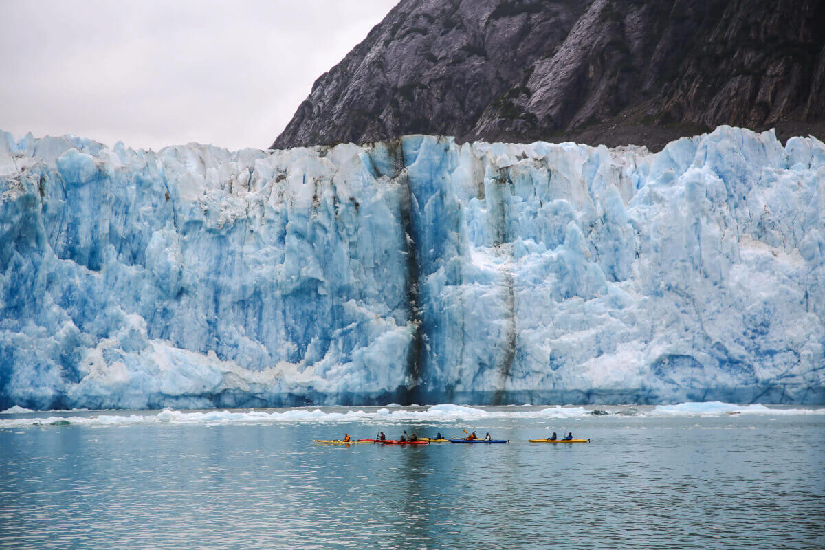 Towering blue glacier meets calm gray water, with tiny kayaks below and dark mountain cliffs behind, creating a dramatic scene.