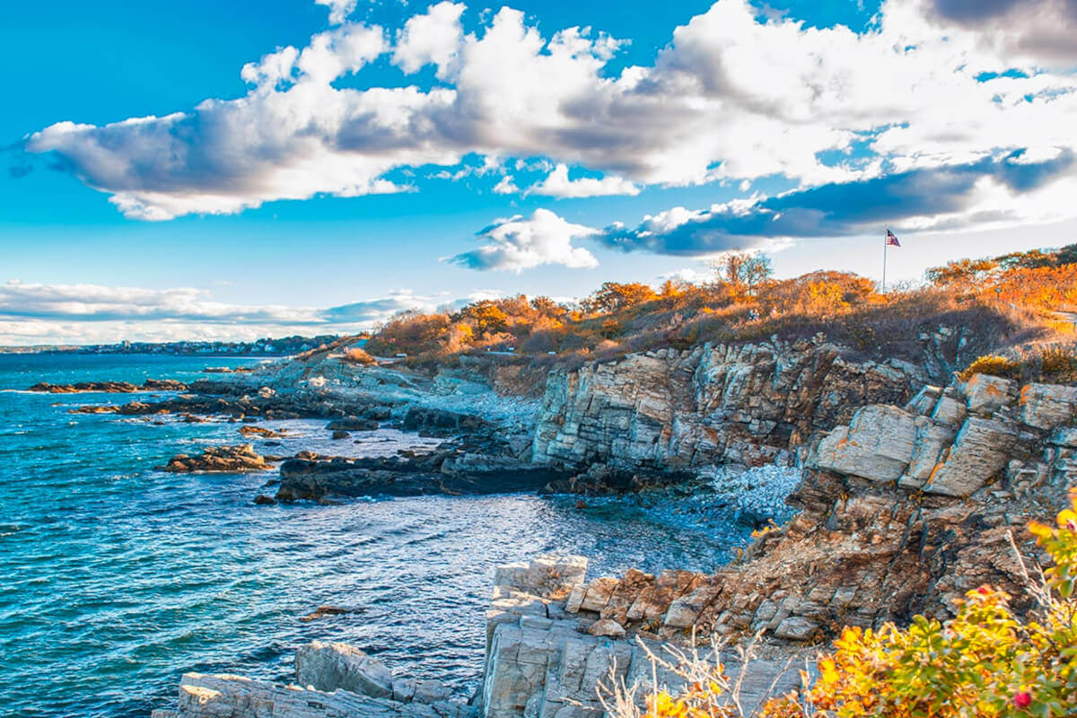 A rocky coastline with yellow-leafed bushes and trees, and an american flag in the distance.