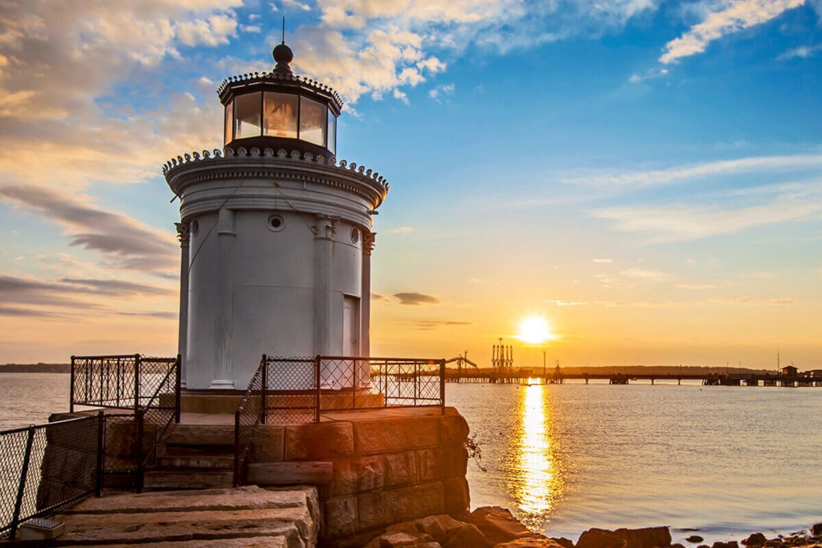 A lighthouse stands along the coastline with the sun rising in the east.