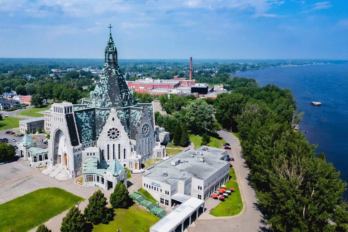 Aerial view of a Gothic Revival church beside a blue lake, surrounded by trees, roads, and buildings under a bright summer sky.